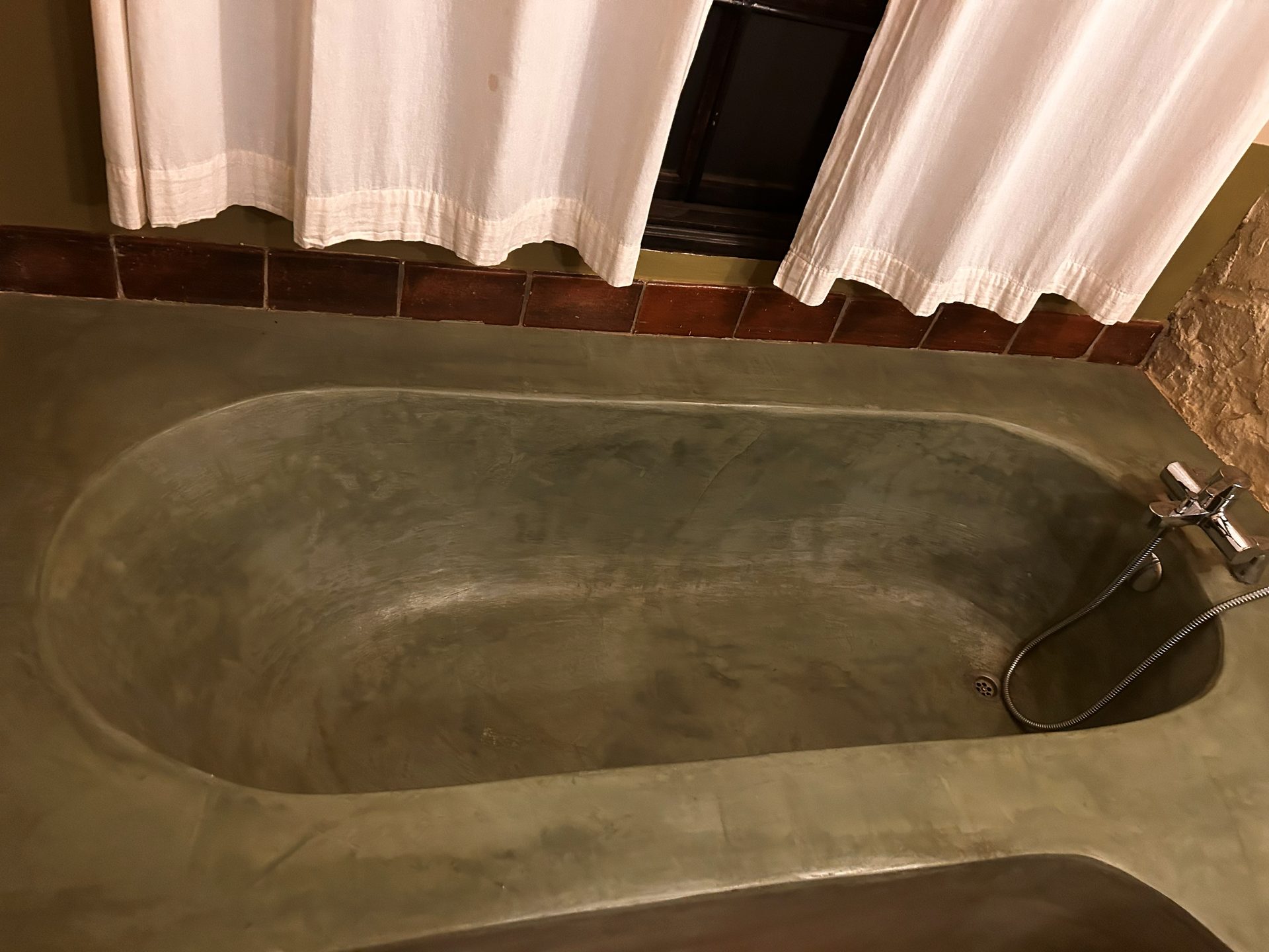 The Rivertrees soaking tub — smooth grey concrete, terracotta tile surround, white curtain, exposed stone wall